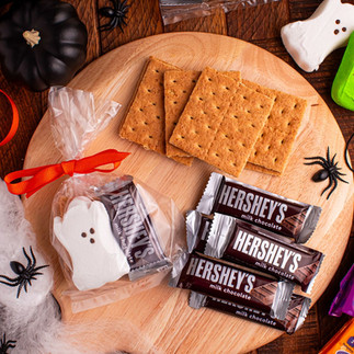Festive Halloween s’mores setup on a wooden board featuring graham crackers, Hershey’s chocolate bars, and ghost- and Frankenstein-shaped marshmallows. Surrounded by black and white mini pumpkins, fake spiders, cobwebs, and colorful candy packaging, creating a cozy and playful fall treat display.