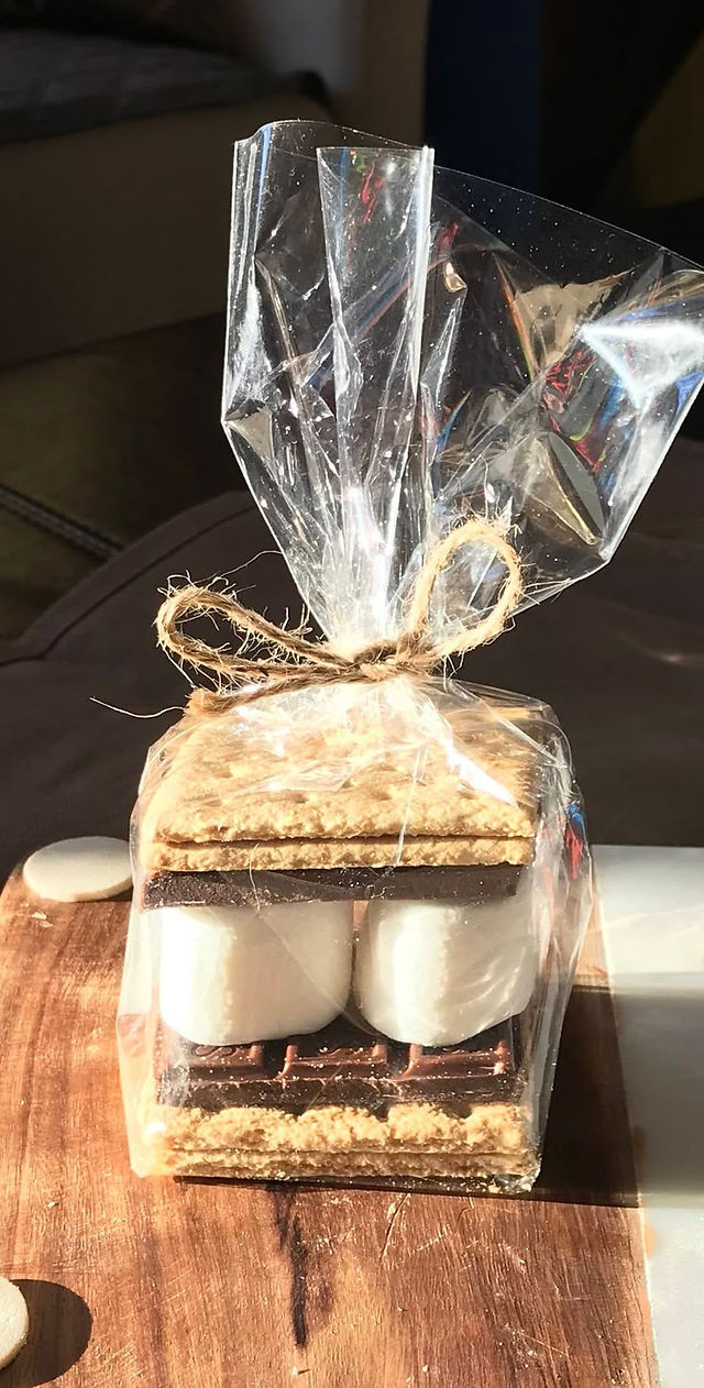 Individually packaged s'mores kit featuring graham crackers, a chocolate bar, and marshmallows stacked neatly in a clear cellophane bag, tied with a rustic twine bow. The kit is placed on a wooden surface, ready to be given as a summer treat or party favor.
