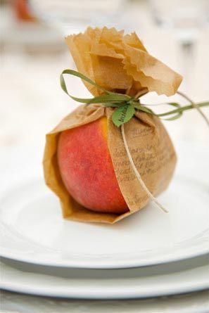 A single peach wrapped in brown parchment paper and burlap, tied with twine and a green ribbon, sitting atop a white plate. The natural packaging gives a rustic, elegant look—ideal for a summer wedding favor or farm-to-table themed event.