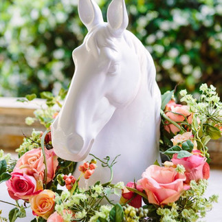 White ceramic horse head centerpiece surrounded by a floral wreath of pink and peach roses with greenery, set outdoors with a blurred garden background.