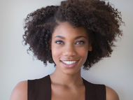 Jeune femme souriante avec des cheveux bouclés, portant une robe noire. Fond gris clair, ambiance chaleureuse et accueillante.