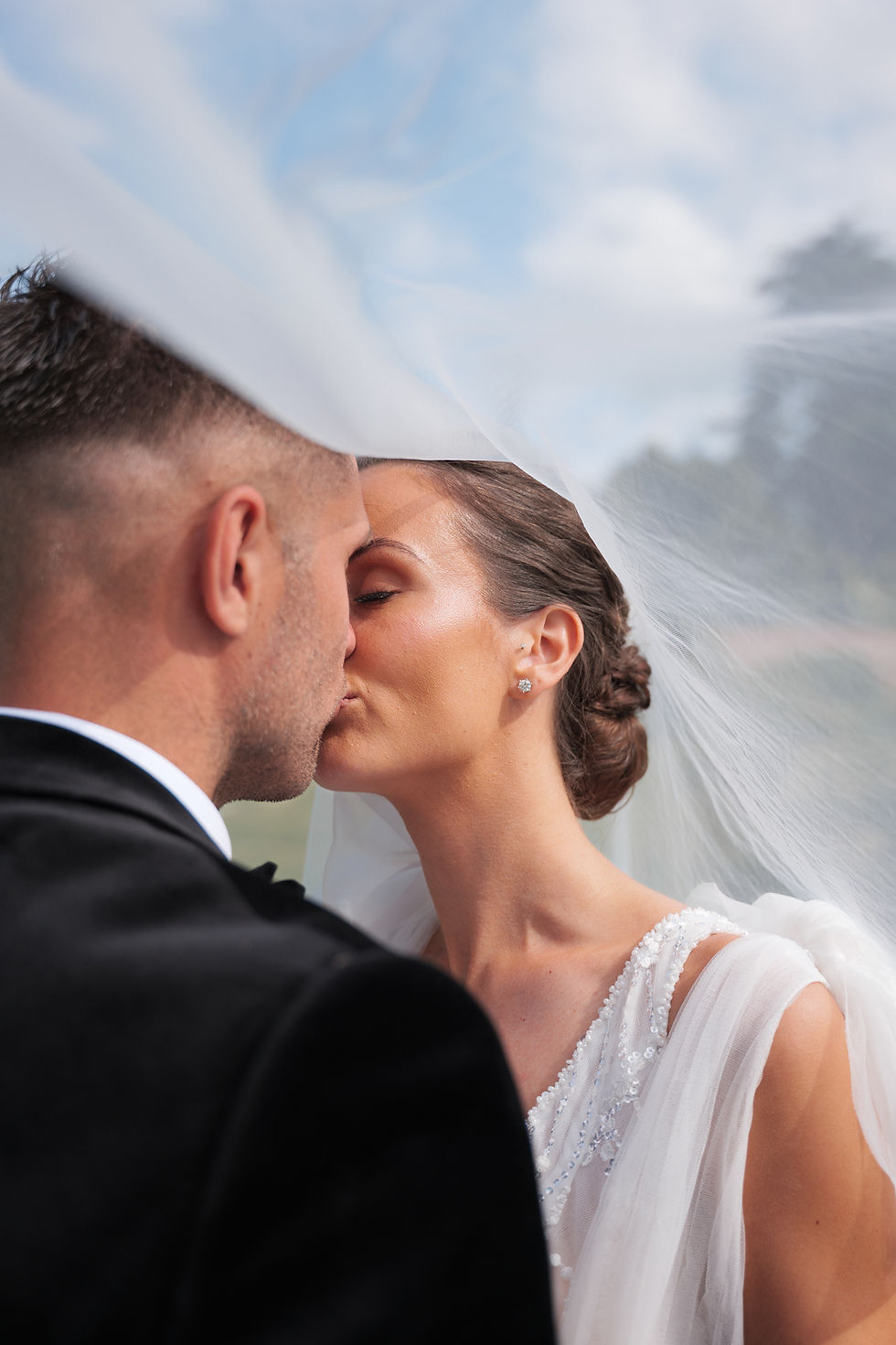 Bride and groom kiss under a sheer veil. The bride wears a beaded dress. Soft lighting and blue sky create a romantic mood.