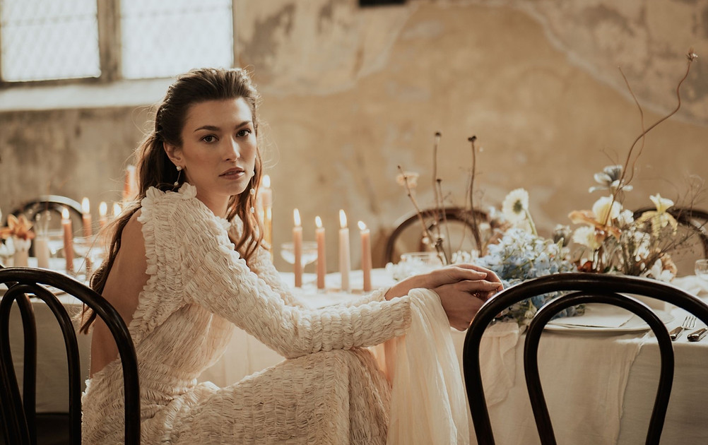 Woman in wedding dress sits at table with flowers. Fineshade Studios.