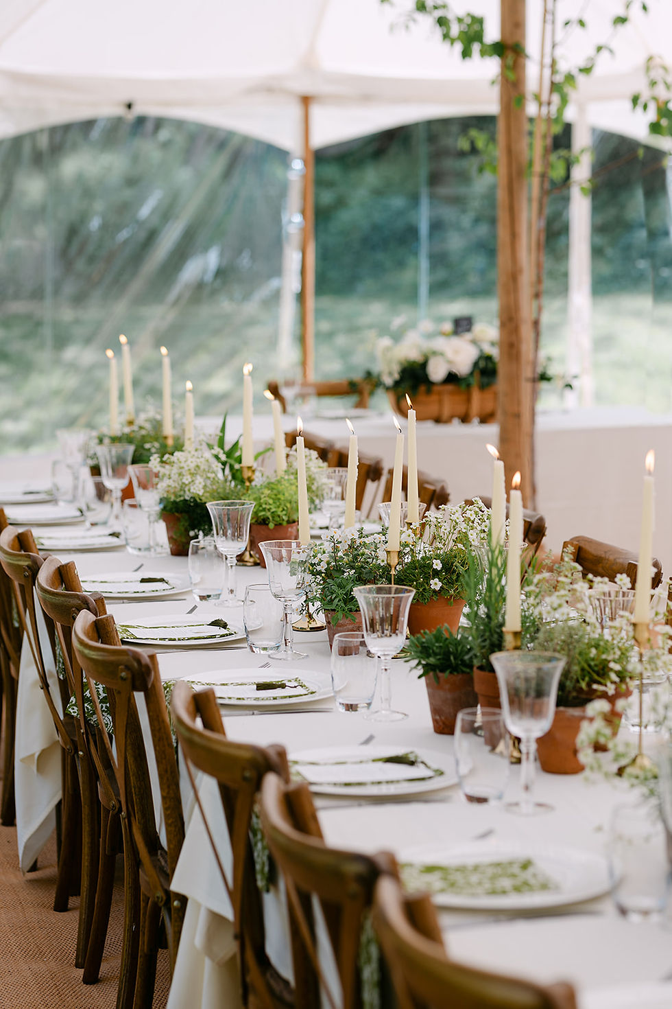 Elegant dining table under a tent with wooden chairs, white tablecloths, tall lit candles, and green floral centerpieces, creating a serene setting.