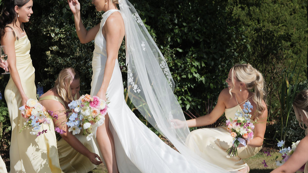 Four people posing for a wedding photo: two men in suits, two women in dresses holding bouquets. They're on grass with trees in the background.