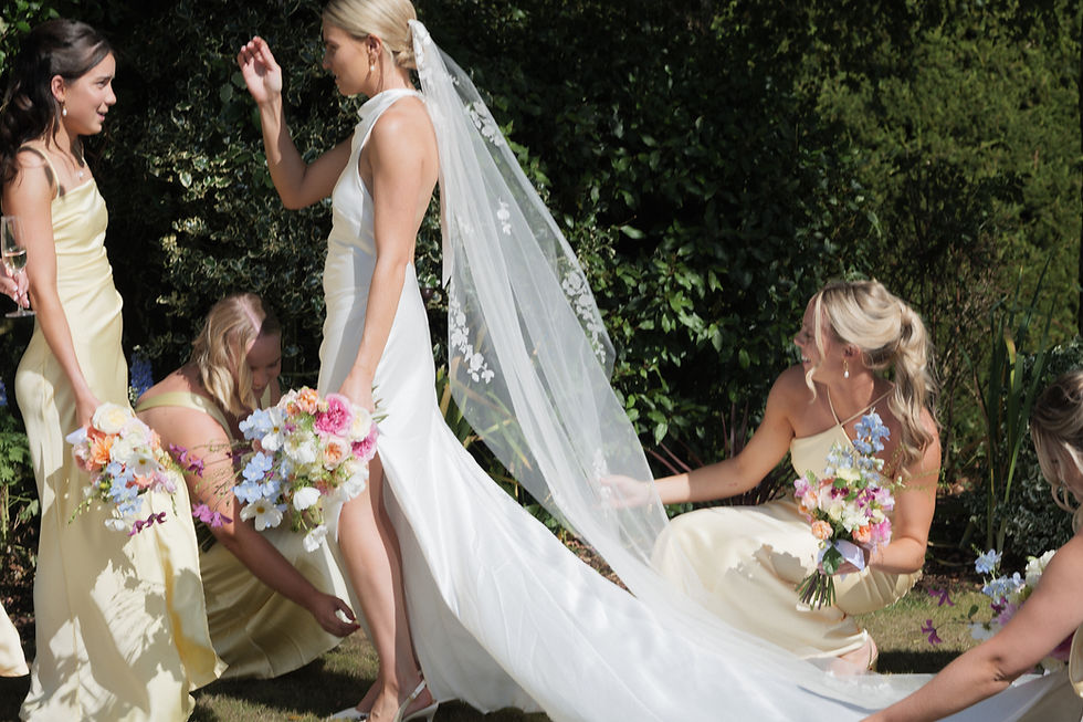 Four people posing for a wedding photo: two men in suits, two women in dresses holding bouquets. They're on grass with trees in the background.