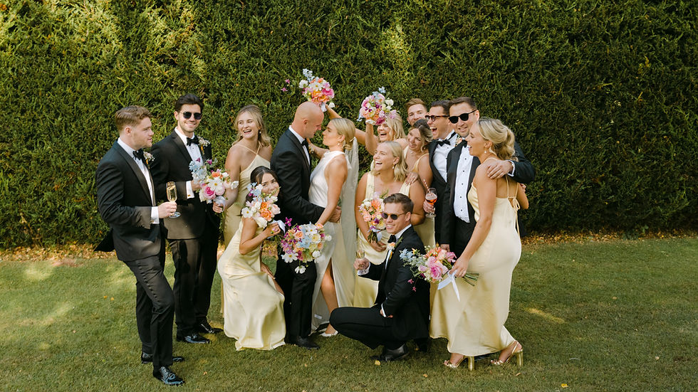 Four men in tuxedos hold a smiling bride in a white dress outdoors. The background features lush green fields under a clear sky.