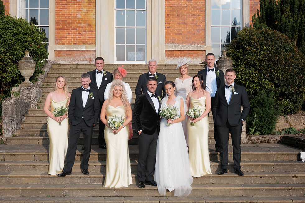 Wedding group poses on stone steps, with bridesmaids in cream dresses and groomsmen in tuxedos. Brick building and greenery in background.