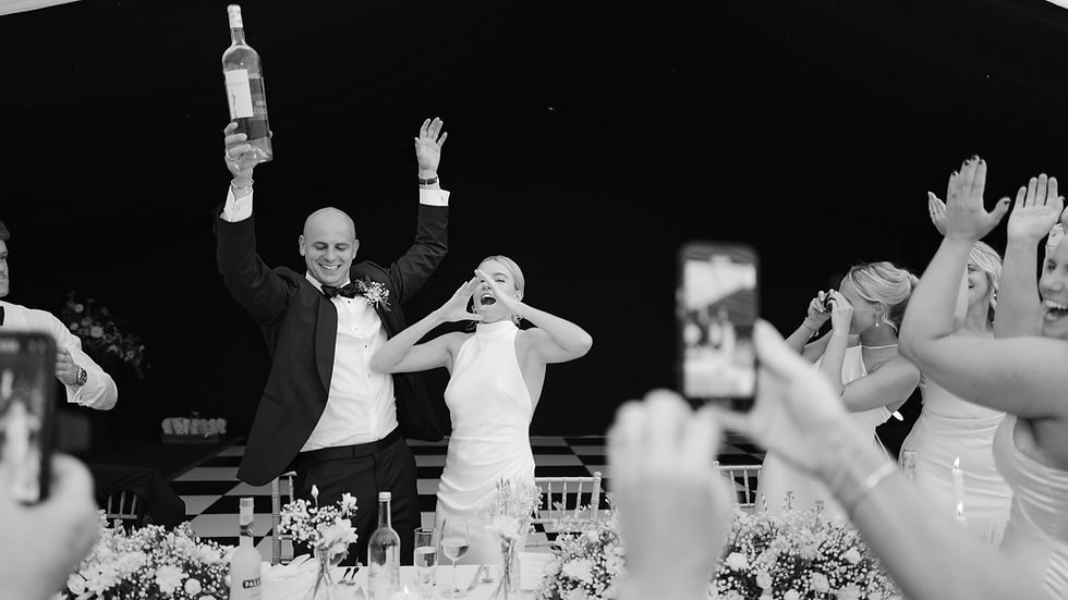 Bride and groom celebrate with a large wine bottle, surrounded by guests in a festive setting. Joyful mood, floral decorations, people cheering.