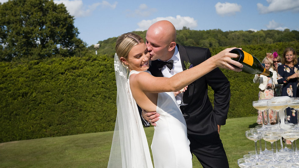 A smiling couple cuts a tiered wedding cake on a wooden barrel. The woman wears a white dress, and the man is in a suit. Black and white image.