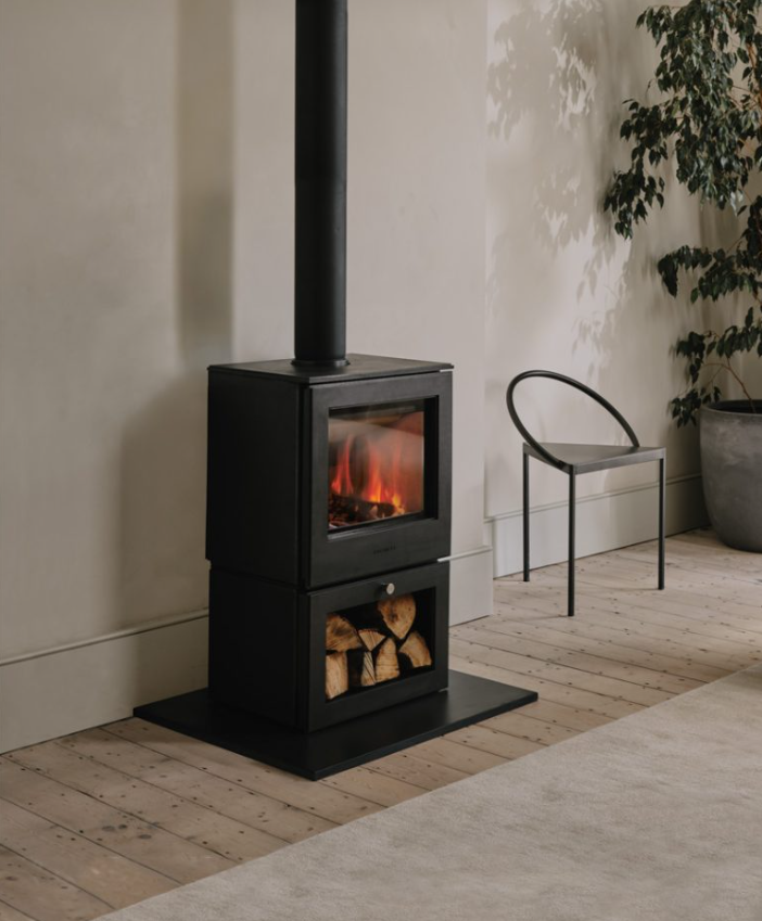 Black Chesneys Serendipity wood-burning stove with visible fire, logs stored below. Located on wooden floor, adjacent to a modern chair and potted plant. Cozy mood.