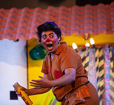 Foto do espetáculo "Las Manas à Obra". Na imagem vemos uma palhaça segurando um veneno e espantada.