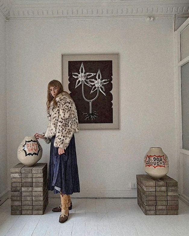 female artist in a fur coat in an exhibition space with big vases and a big painting with metallic flowers on a brown surface