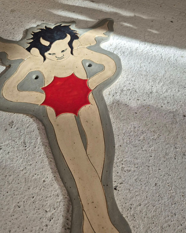 Closeup of a stoneware wallhanging in grey, beige, depicting a devilish angel holding a bursting red star