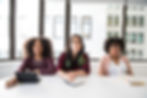 Three women sitting at a desk. looking up at someone off camera.