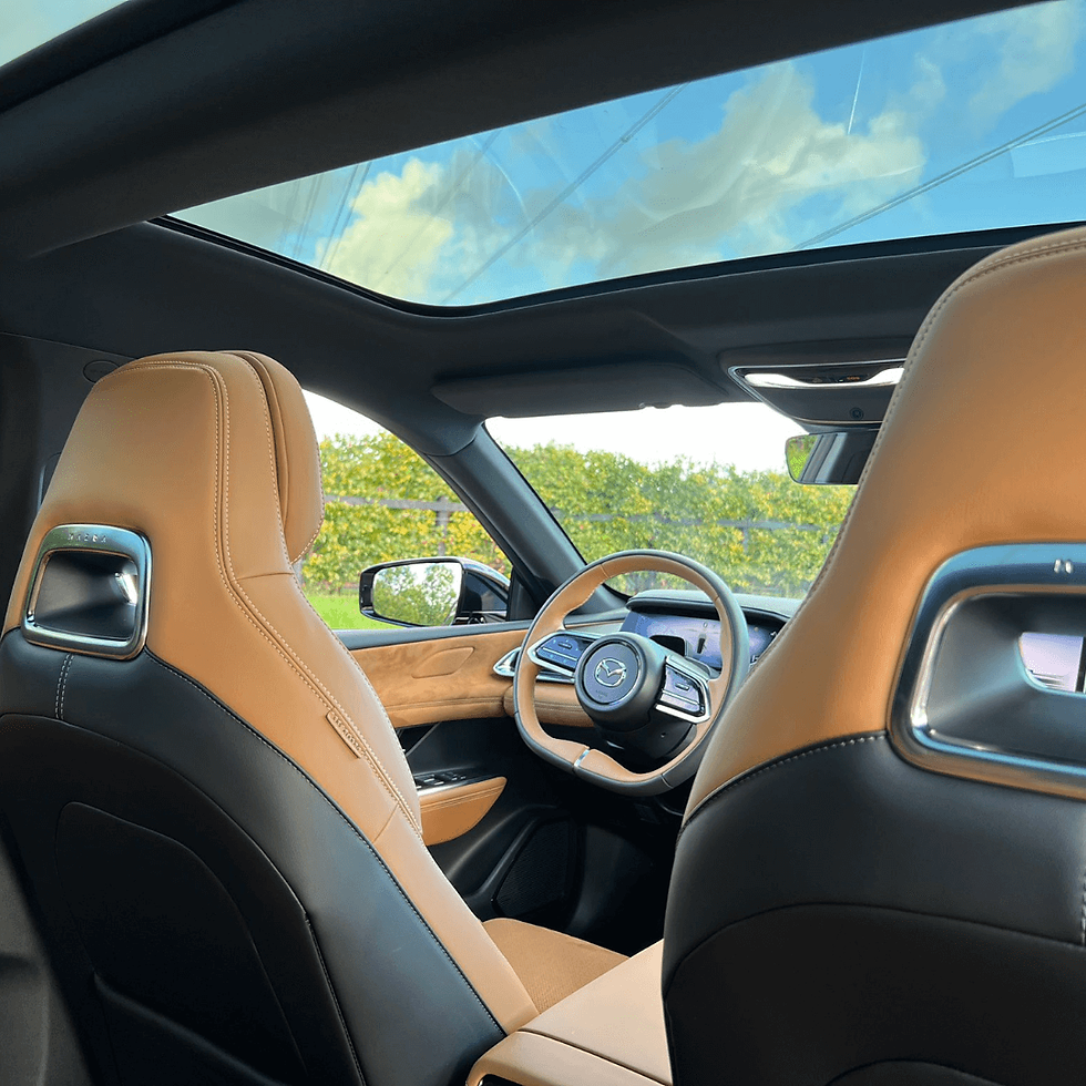 Car interior with tan leather seats, panoramic sunroof, and steering wheel featuring a Mazda logo. Blue sky and trees visible outside.