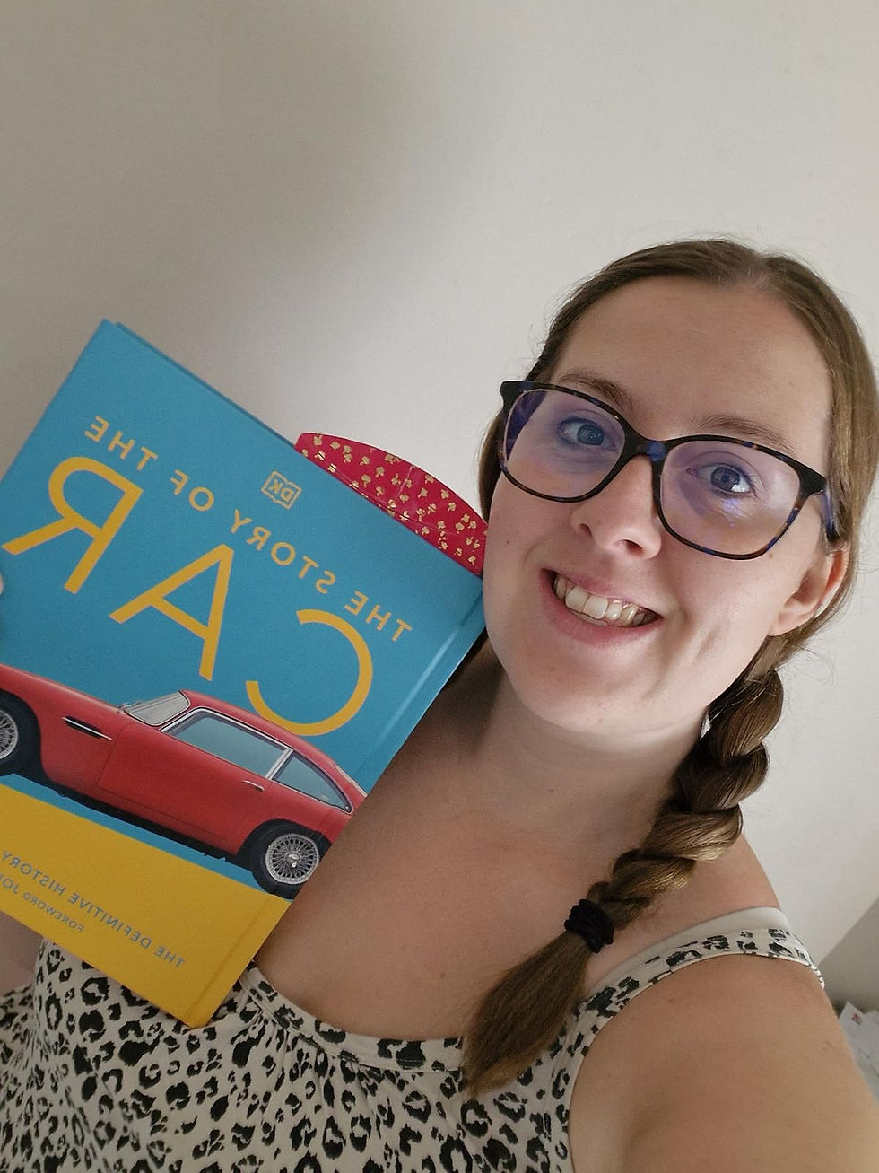 Woman with glasses smiles, holding "The Story of the Car" book. Bright blue cover with red car image. Leopard-print top. Plain background.