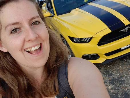 Smiling woman taking selfie in front of a yellow Ford Mustang with black stripes. Sunny day, parked cars in the background.
