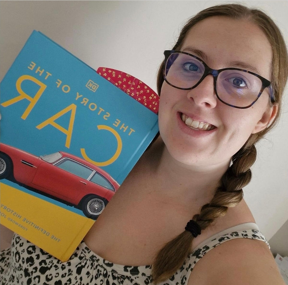Smiling woman with glasses holds a blue book titled "The Story of the Car" featuring a red car. She wears a patterned top.
