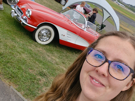 Woman in glasses smiles in front of a vintage red and white convertible car. Two people stand by a tent in the grassy background.