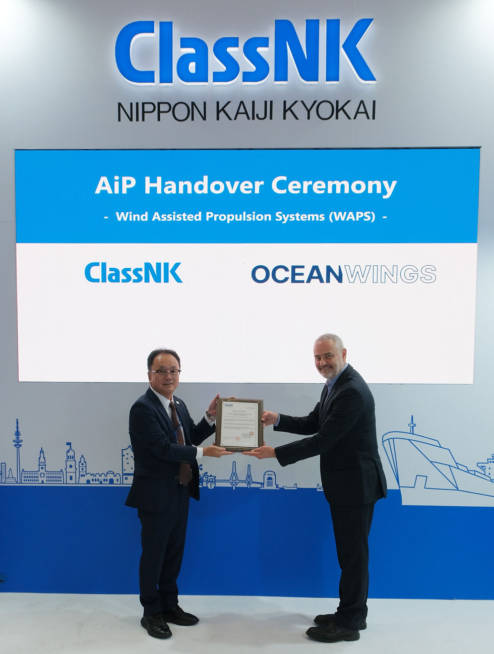 Two men in suits exchange a framed certificate at ClassNK’s AiP Handover Ceremony for OceanWings WAPS.