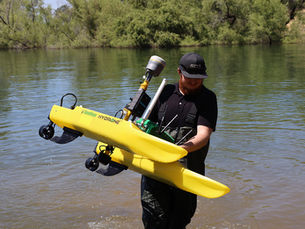 Man in waders carries a yellow Seafloor HYDRONE from a lake with wooded shoreline. He's wearing a cap and appears focused.