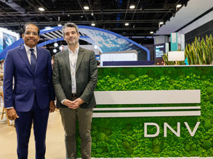 Two men in suits stand smiling next to a green, plant-covered booth labeled DNV. Background shows a modern conference setting.