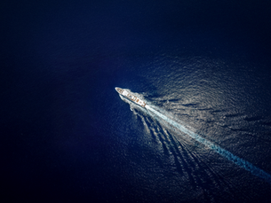 Aerial view of a white yacht sailing on dark blue ocean, leaving a foamy wake. The scene reflects tranquility and motion.