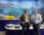 Two men on an exhibition stand with a maritime scene as a backdrop