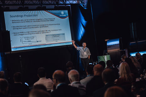 A man presenting stage in a blue shirt with a large projector screen behind him