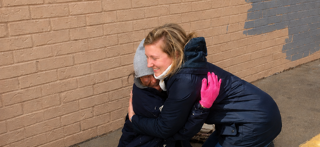 A woman embraces another person beside a brick wall, offering comfort and encouragement during community outreach.