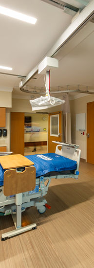 Kathryn Kirk Center designed by Architectural Nexus. Photo shows the interior view of a medical room with a hospital bed in the center. Facing the bed is the custom full wall millwork with storage cabinets and a wall mounted large screen TV. White ceilings with both thin rectangular lights and recessed round light fixtures that provides great illumination to the room. Light wood paneling flooring are bordered by light beige painted walls. Next to the entry door is a countertop with a sink and cabinets above and below the counter. The room walls and ceiling shows face mounted top of the line medical equipment. Next to the bed is a side table with medical equipment and a telephone.