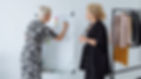 Two women in a bright studio plan fashion looks at a flip chart, with a clothing rack of jackets and skirts beside them.