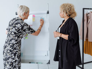 Two women in a bright studio plan fashion looks at a flip chart, with a clothing rack of jackets and skirts beside them.