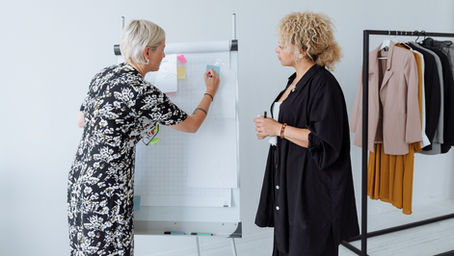 Two women in a bright studio plan fashion looks at a flip chart, with a clothing rack of jackets and skirts beside them.