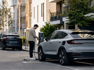 Man charging a silver electric SUV at a curbside station on a quiet city street, with a black car and apartments behind.