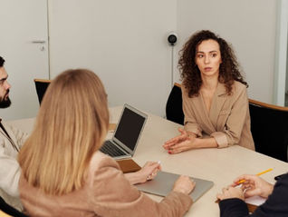 Business meeting with a woman speaking at a table, others listening շուրջ a laptop and notebook in a plain office.