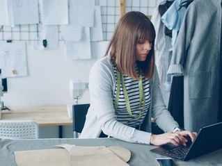 Woman seamstress works on a laptop in a clothing studio, with sewing patterns, mannequins, and a focused mood.