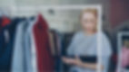 Blonde woman in a clothing store studies a tablet beside racks of hanging clothes, focused and calm.