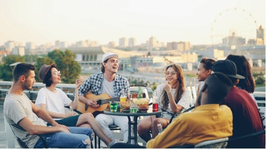 Friends sit on a rooftop patio, one strumming a ukulele, sharing drinks and laughing with city skyline and Ferris wheel behind.