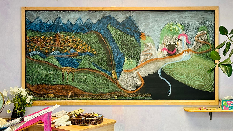 Colorful chalk drawing on blackboard showing mountains, river, village, and tunnel. Books and flowers on a table below. Calm and creative atmosphere.
