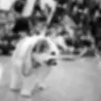 Black and white photography of a scruffy dog with a bandana playfully holding its own leash in its mouth. The background is softly blurred, showing a group of people in what appears to be a classroom or public space.