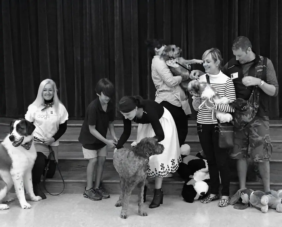 Black and white candid photography of a diverse group of people petting and interacting with multiple dogs indoors. The scene appears warm and communal, likely a volunteer animal event or gathering.