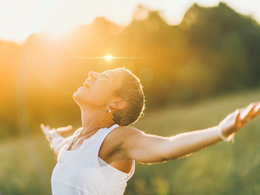 A woman stands with her arms wide and her eyes closed in a sunny feild. 
