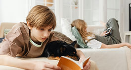 Boy reads book next to black dog with sister sat behind on other sofa