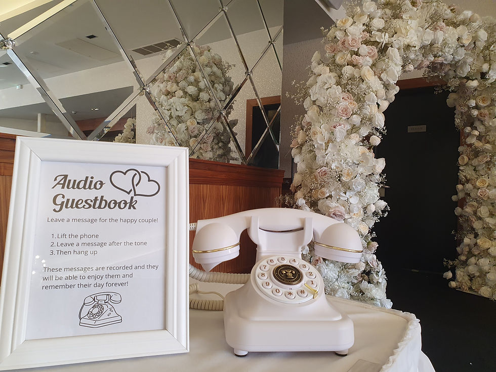 A white audio guestbook phone sat next to a white framed sign in front of a arch of white flowers.