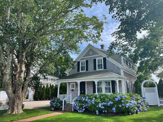a quaint two-story house with gray shaker shingles and white trim surrounded by bright green trees - Bright