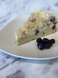 Slice of blueberry cream cheese coffee cake on a white plate and a pile of blueberries next to the cake slice. On a grey and white marble background.