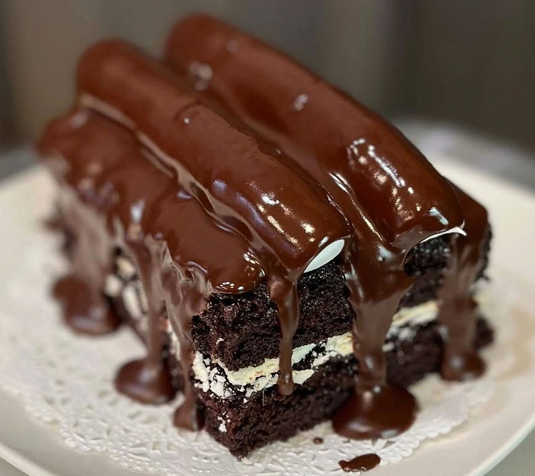 Slice of Bumpy-style cake. Double layer chocolate cake with buttercream filling topped with two pipes of buttercream and rich chocolate ganache dripping down over the cake and onto a white doily.