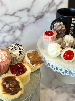 A galvanized metal tray on the bottom left holds our strawberry, oreo, apple crumble, cherry fruit topped, and turtle Original NY Style Cheesecake cupcakes in two rows. Next to a white pedestal stand with strawberry, lemon, chocolate, and cherry topped Original NY Style Cheesecake bite-sized tartlets. A black cup labled coffee in white font is behind the chocolate tartlet all on a white and grey marble background. 
